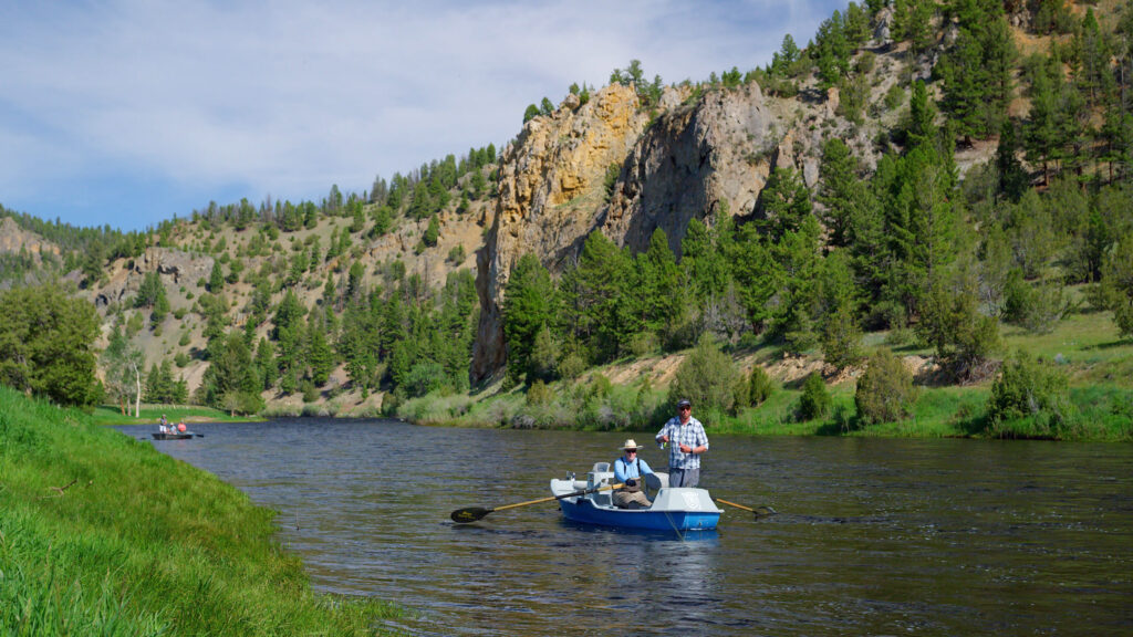 A drift boat carries two fly fishermen down the Big Hole River near Wisdom and Twin Bridges, Montana, with steep pine-covered cliffs rising above the water and another boat floating in the distance. Photo by Tempest Technologies.