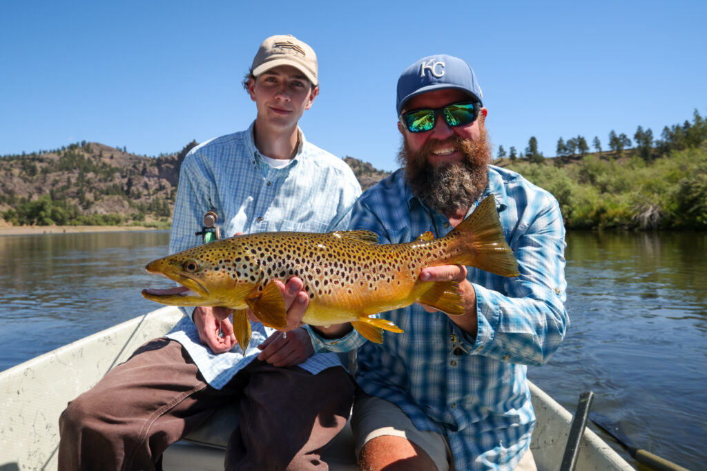Two anglers aboard a drift boat hold a large, heavily spotted brown trout caught on the blue ribbon section of the Missouri River near Craig, Montana, with grassy bluffs lining the riverbank behind them. Photo by Sarah Bolt, Tempest Technologies.