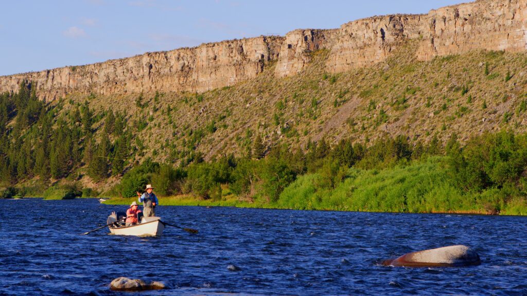 An angler casts a fly rod from the bow of a drift boat on the Madison River near Ennis, Montana, floating past sagebrush-covered cliffs as a guide rows downstream in the evening light. Photo by Mike Dreesman, Tempest Technologies.