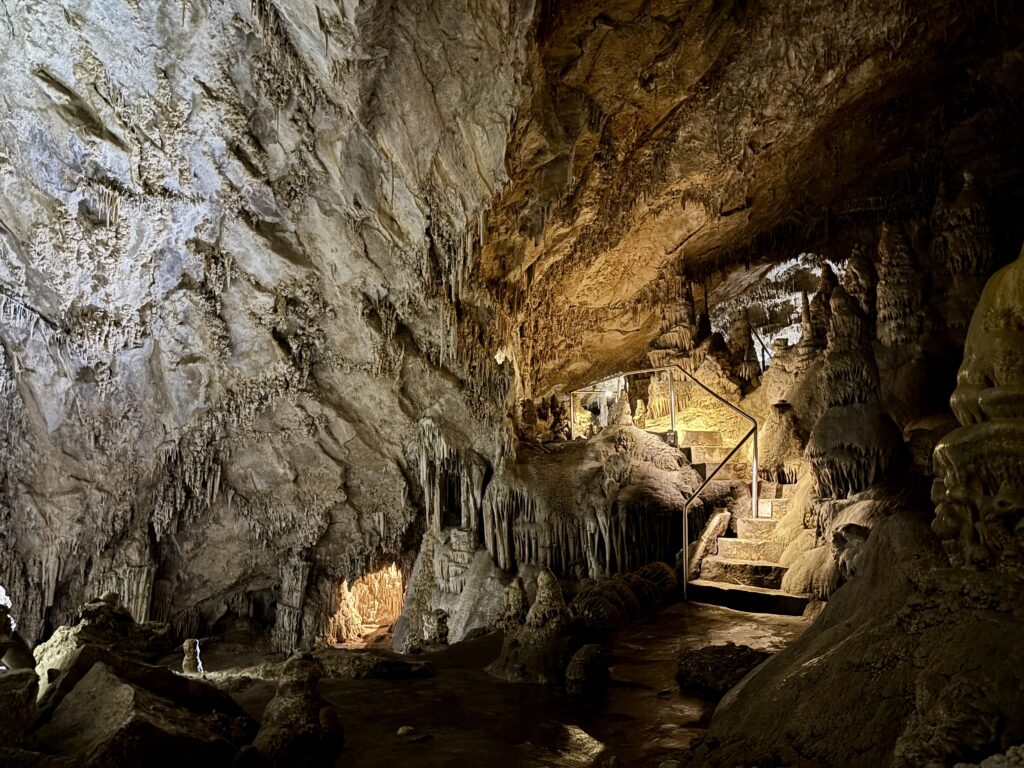 A staircase with handrails winds through an illuminated chamber on the Classic Tour at Lewis and Clark Caverns State Park, surrounded by dramatic stalactite and flowstone formations glowing gold and grey under the cave's lighting.