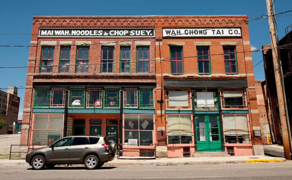 Exterior of the Mai Wah Museum in Butte, Montana — two connected two-story brick buildings with green and salmon-painted trim. Bold painted signage reads "Mai Wah, Noodles & Chop Suey" and "Wah Chong Tai Co." against a bright blue sky. A dark SUV is parked along the street in front. Photo by Donnie Sexton, Montana Dept. of Tourism.