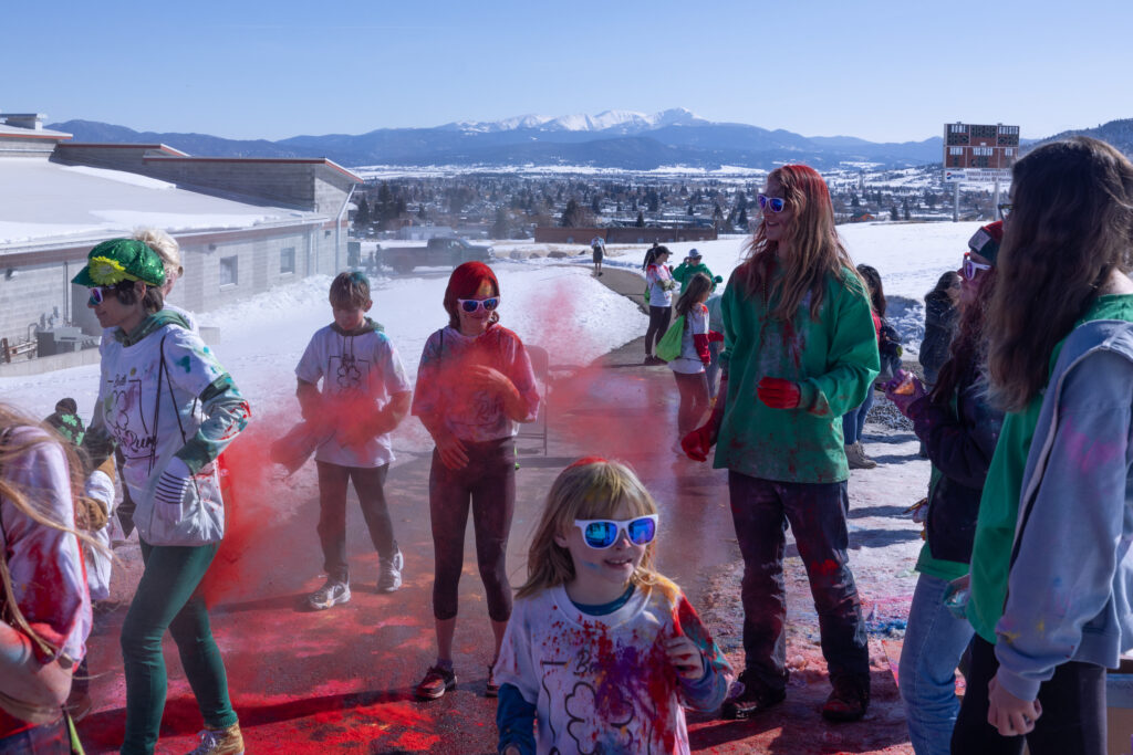 Kids and adults laugh and toss red color powder during the Butte Cares St. Patrick's Day Color Run, with snow-covered mountains visible in the background. Family friendly St. Patrick's Day activity in Uptown Butte, Montana. Photo by Sarah Bolt, Tempest Technologies. 