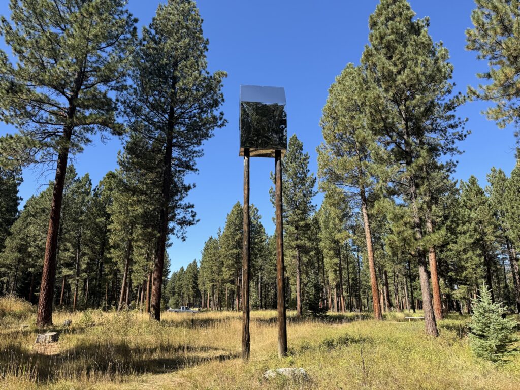House of Sky by Alan Counihan, a mirrored, house-shaped box perched high atop two tall wooden poles, reflects the surrounding pines and blue sky above a golden, grassy clearing at Sculpture in the Wild in Lincoln, Montana. Photo by Alyssa Starr, Tempest Technologies, LLC.