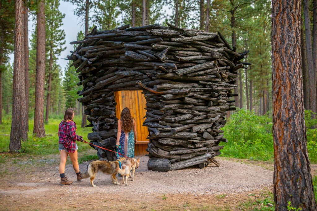 Two visitors walk a pair of leashed, tan and gray dogs along a gravel path toward a rounded sculpture of stacked, charred logs at Sculpture in the Wild in Lincoln, Montana, its doorway opening to a warm, golden wood-paneled interior with a built-in bench, surrounded by tall pines and green undergrowth. Photo by Eric Bunting.