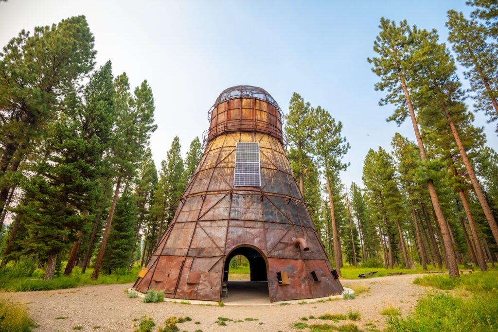 Re-Imagining the Delaney Sawmill TeePee Burner by Kevin O'Dwyer, a 75-foot cone of rust-colored steel topped with a domed mesh cap, rises above a gravel path at Sculpture in the Wild in Lincoln, Montana, with a solar panel on its side that powers its nighttime glow and an arched doorway at the base opening into the interior, surrounded by tall pines. Photo by Eric Bunting. 