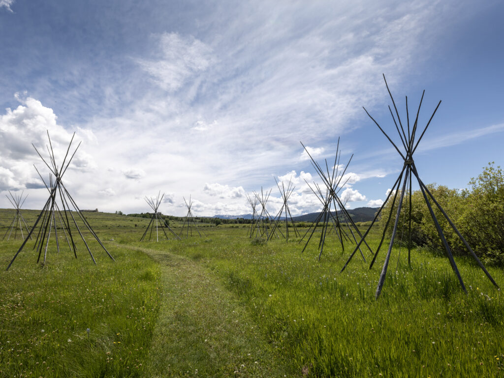 Wooden tipi frames stand in a wide green meadow under a dramatic blue sky at Big Hole National Battlefield near Wisdom, Montana in Southwest Montana, marking the historic Nez Perce camp.
