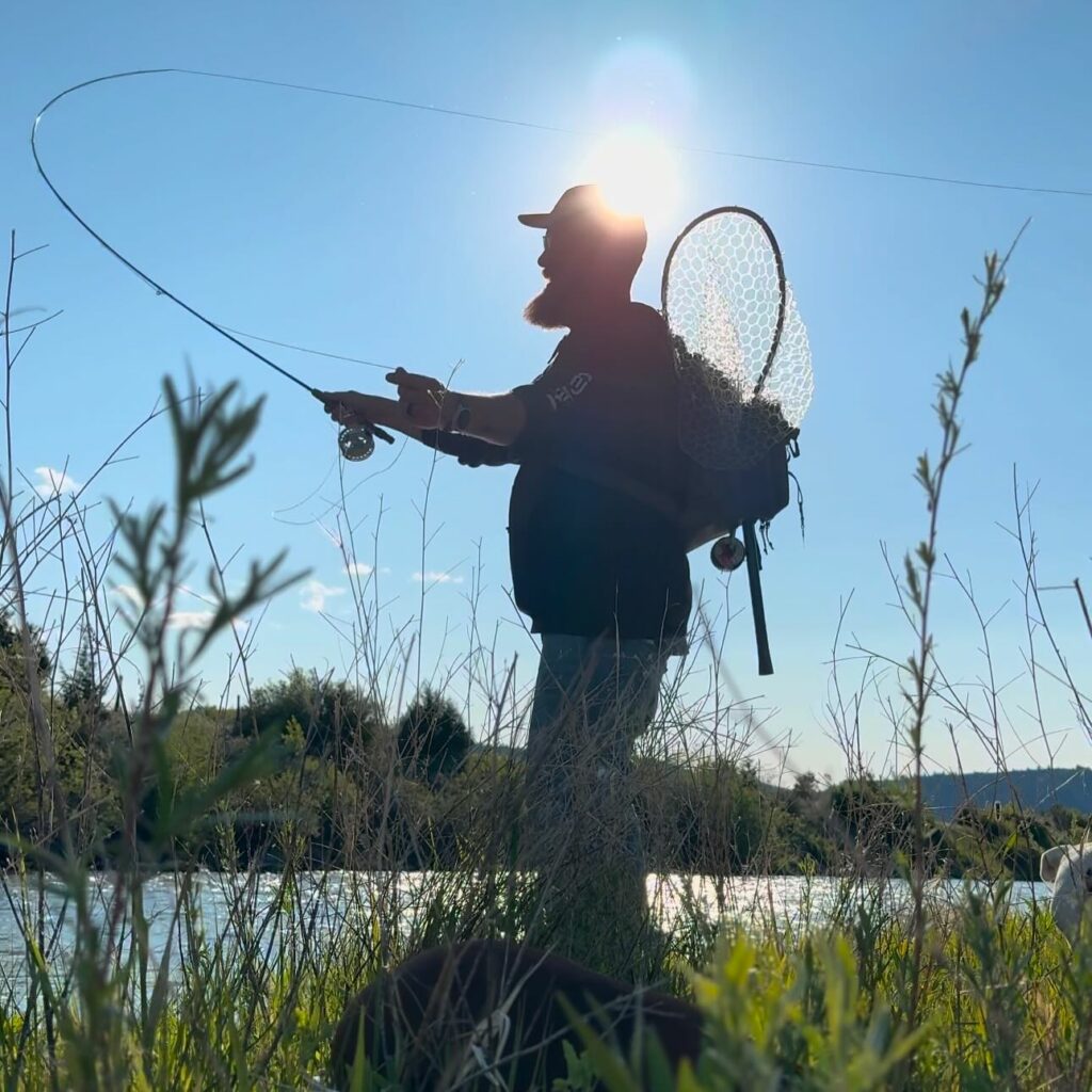A fly fisherman casts a fly rod on the Big Hole River in Southwest Montana, a blue-ribbon trout stream known for its rainbow and brown trout populations. The angler is backlit by the sun with riverbank grasses in the foreground and a landing net on his back. Photo by @_0ffthegrid on Instagram. 