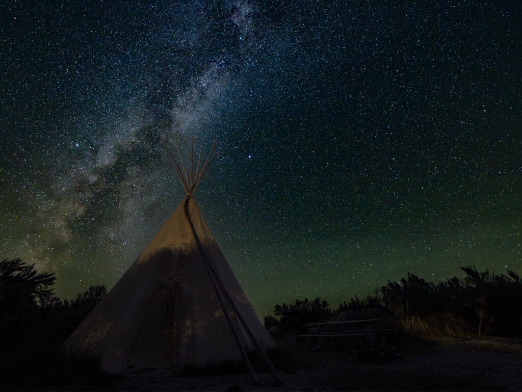 Night sky and Milky Way galaxy photographed above a traditional canvas tipi at Bannack State Park campground in southwest Montana, with thousands of stars and the bright core of the Milky Way arching overhead in vivid blue and teal tones, surrounded by a silhouetted treeline and the dark rolling landscape of Beaverhead County, Montana, offering a glimpse of the immersive stargazing and historic camping experience available at one of Montana's most beloved ghost town state parks