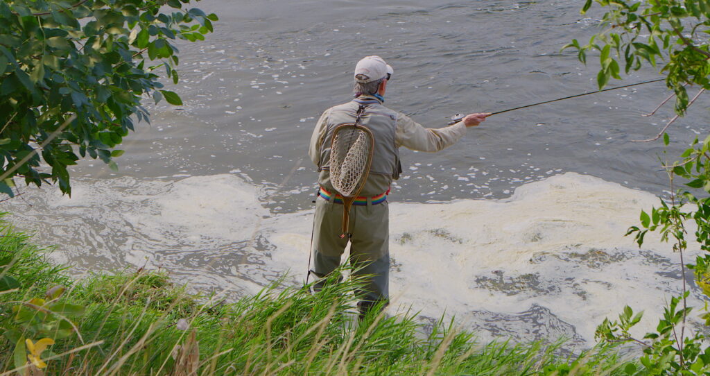 An angler with a net strapped to his back stands at the edge of the Missouri River near Craig, Montana, casting a fly fishing line into the swollen, silty spring waters. Surrounded by green foliage and tall grass, this image captures the beauty and challenge of fly fishing in Southwest Montana during mud season, when high runoff levels make for a strategic and rewarding experience on one of Montana's most iconic rivers. Photo by Mike Dreesman, Tempest Technologies.