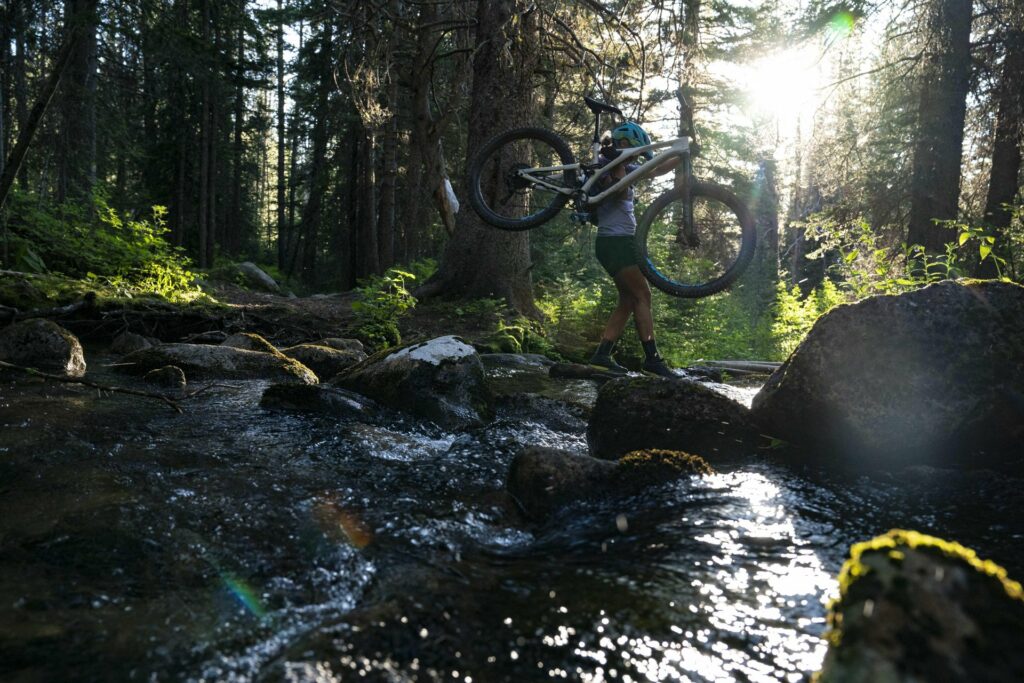A mountain biker in a helmet, shorts, and trail shoes carefully navigates a rocky creek crossing in a dense evergreen forest in Boulder, Montana, hoisting their mountain bike overhead to keep it clear of the fast moving spring water rushing over the moss covered boulders below. Dramatic golden backlight filters through the tree canopy, illuminating the scene and highlighting the adventurous, off the beaten path nature of mountain biking trails in Southwest Montana, where spring runoff adds an extra layer of challenge and beauty to the riding experience. Photo by Garret Smith.
