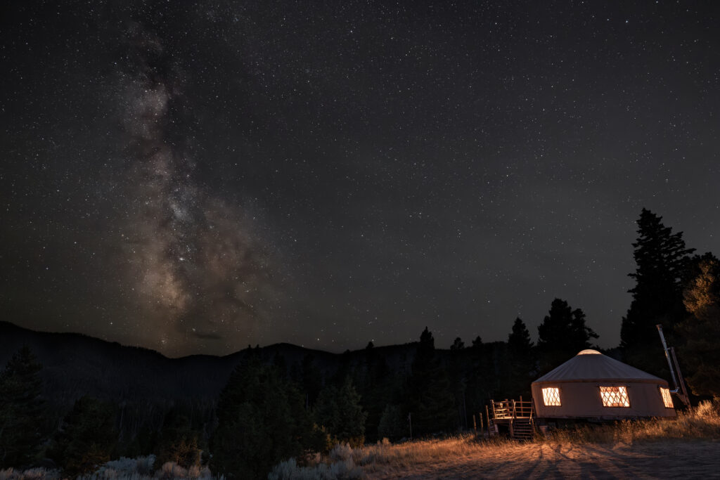 Landscape night sky photograph of the Milky Way galaxy arching over a yurt at Homestake Lodge near Butte, Montana, with a warmly lit yurt glowing from within against the deep darkness of the surrounding landscape, tall silhouetted evergreen trees framing the right side of the frame, rolling foothills and mountains visible in the background beneath a sky dense with stars, highlighting one of Southwest Montana's most unique dark sky lodging, off road ATV and UTV, and Nordic ski destinations in the Deer Lodge National Forest. Photo by Sarah Bolt, Tempest Technologies.