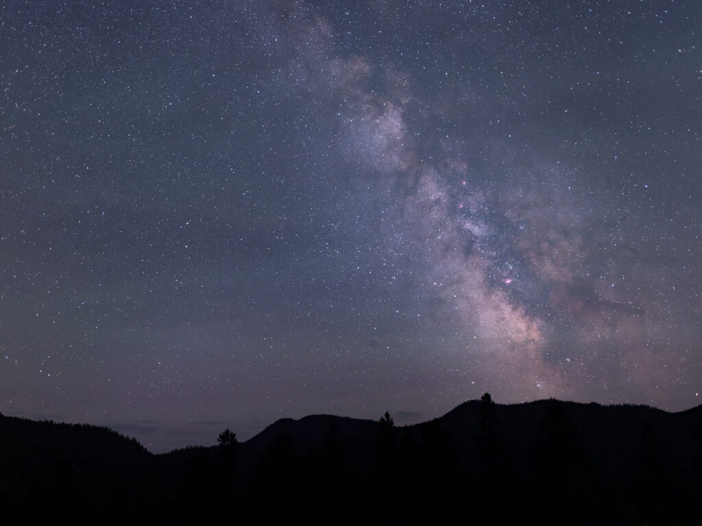 Night sky photograph taken at Homestake Pass near Butte, Montana, looking up at the dense and luminous core of the Milky Way galaxy glowing in shades of purple, blue, and silver against a deeply dark sky, with the bold black silhouette of a mountain ridgeline and scattered pine trees cutting across the lower frame, the faint outline of a tall structure or post visible against the stars, showcasing the exceptional dark sky conditions found along the Continental Divide in southwest Montana just minutes from Butte, Montana. Photo by Sarah Bolt, Tempest Technologies.