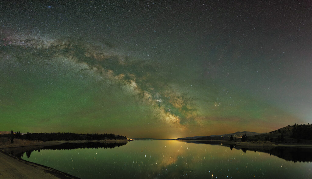 Panoramic night sky photograph of the Milky Way galaxy arching over Canyon Ferry Lake near Helena, Montana, with vivid green and teal aurora-like atmospheric glow illuminating the horizon, golden light reflecting off the calm glassy water below, silhouetted evergreen tree line framing the shoreline, and thousands of stars visible in the dark sky above southwest Montana's most iconic reservoir, taken at the Montana Learning Center, a premier destination for stargazing and astronomy education in Montana.
