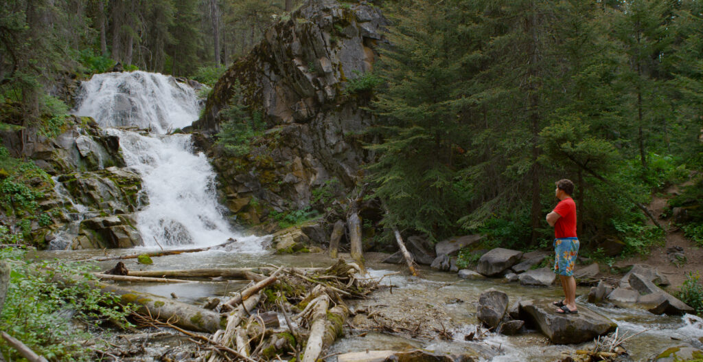 A hiker stands on rocks at the base of Lost Creek Falls near Anaconda, Montana, gazing up at the rushing white waterfall as it tumbles down a jagged granite cliff face into a rocky creek below. Fallen logs, moss covered boulders, and towering evergreen trees frame the scene at Lost Creek State Park, one of the most accessible and stunning waterfall destinations in Southwest Montana. Photo by Mike Dreesman, Tempest Technologies.
