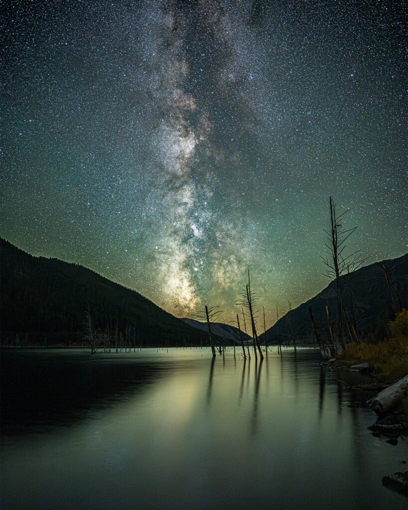 Vertical night sky photograph taken at Earthquake Lake in southwest Montana's Madison River Valley, capturing the brilliant core of the Milky Way galaxy arching over a dramatic mountain ridgeline, with the eerie silhouettes of standing dead trees rising from the still reflective water below, a haunting and beautiful reminder of the 1959 Hebgen Lake earthquake that created this unique geological landmark, showcasing southwest Montana's extraordinary dark sky photography locations and the otherworldly landscape of the Lee Metcalf Wilderness corridor near West Yellowstone. Photo by @fatkitty13 on Instagram. 