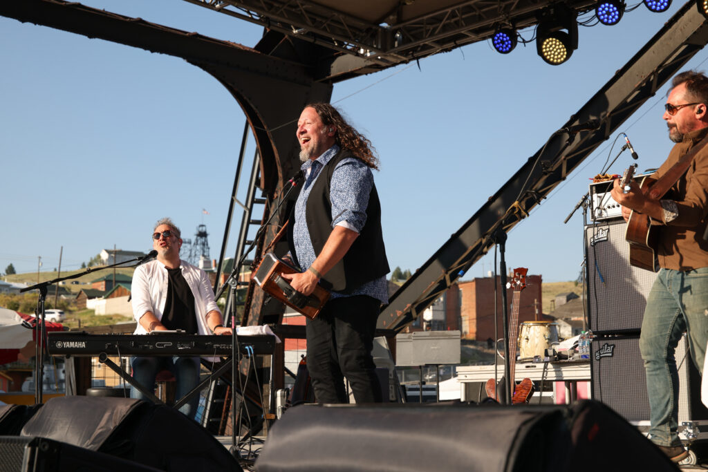 Three musicians perform on an outdoor stage at the Montana Folk Festival in Uptown Butte, Montana, with the center vocalist singing and holding a guitar under a clear blue sky, historic mine headframes and Butte's brick buildings framing the scene behind the stage. Photo by Sarah Bolt, Tempest Technologies.
