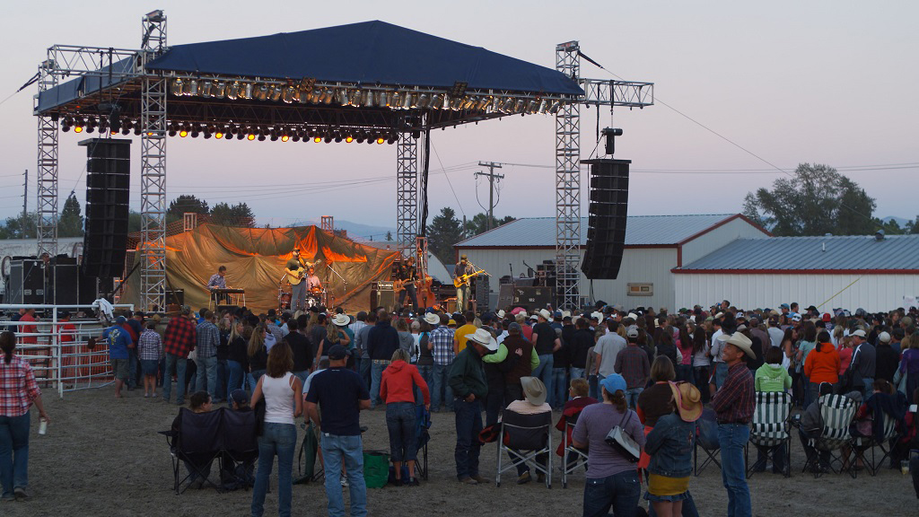 Live concert on the dirt grounds of the Dillon rodeo at Montana's Biggest Weekend, with a large crowd of western-dressed attendees watching a band perform on a floodlit outdoor stage as the sun sets behind the mountains. Photo by Mike Dreesman, Tempest Technologies.