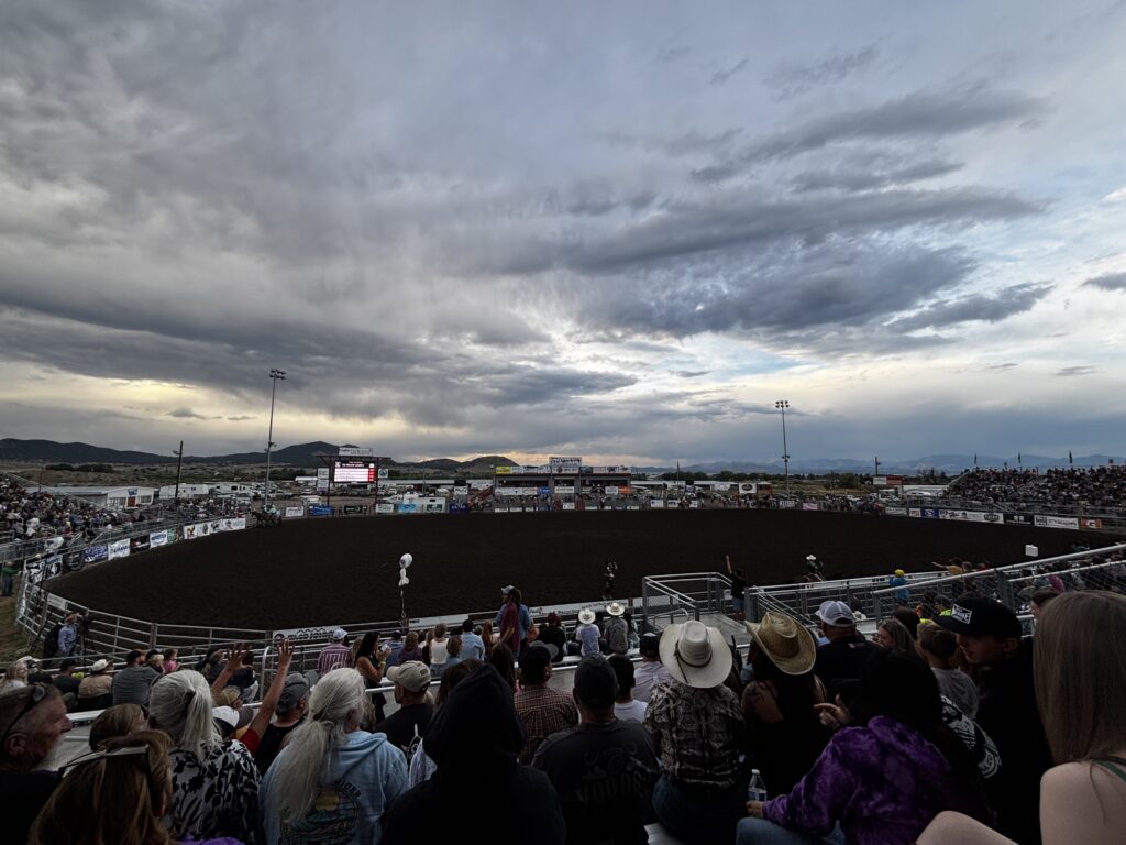 The rodeo arena at the Last Chance Stampede at the Lewis and Clark County Fairgrounds in Helena, Montana, filled with spectators in the grandstands as competitors prepare in the dirt arena below, with a stormy evening sky and the Helena valley's mountain silhouette stretching across the horizon