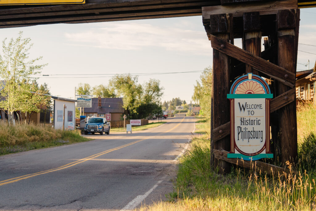 The iconic 'Welcome to Historic Philipsburg' entry sign, featuring teal and red decorative detailing mounted within a heavy weathered timber frame, stands roadside as vehicles pass along the main highway into Philipsburg, Montana, with local businesses, green summer trees, and a pale blue sky stretching down the two-lane road into this charming Flint Creek Valley community in southwest Montana. Photo provided by Trout Unlimited