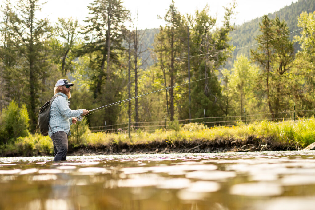 A fly fisherman wades knee-deep in a clear, sun-dappled river near Philipsburg, Montana, mid-cast with his fly line arcing gracefully through the air above the water, surrounded by green summer foliage, tall conifers, and the forested slopes of the Flint Creek Valley, capturing the world-class Blue Ribbon fishing experience that helped earn Philipsburg its designation as Trout Unlimited's inaugural Conservation Town Champion. Photo provided by Trout Unlimited.