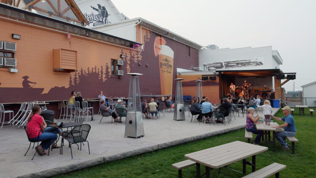 Here are a few options: "Guests enjoy craft beer and live music on the outdoor patio stage at Lewis and Clark Taproom in Helena, Montana, with patio heaters, string lights, and a large beer mural decorating the brewery's exterior wall on a summer evening" "A band performs on the covered outdoor stage at Lewis and Clark Taproom in Helena, Montana, as patrons relax at patio tables and picnic benches with craft beers in hand beneath a large painted mural on the brewery wall. Photo by Mike Dreesman, Tempest Technologies.