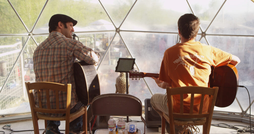 Two acoustic guitarists perform back to back inside the iconic geodesic dome at Norris Hot Springs in Norris, Montana, with the misty outdoor pool and surrounding trees visible through the transparent dome walls behind them. Photo by Mike Dreesman, Tempest Technologies.