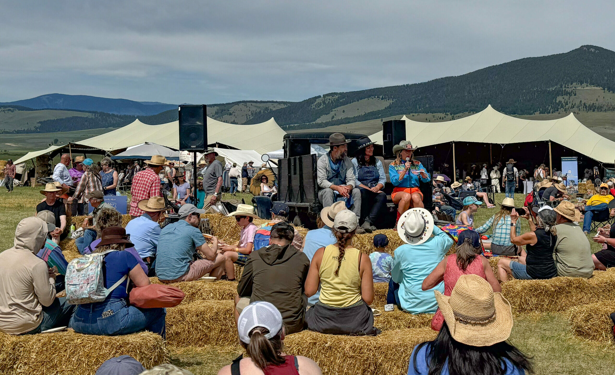 Festival attendees on hay bales facing an outdoor stage at the Old Salt Festival in Helmville, Montana, with the Blackfoot Valley's open ranchland and mountains in the background. Photo by Tempest Technologies.