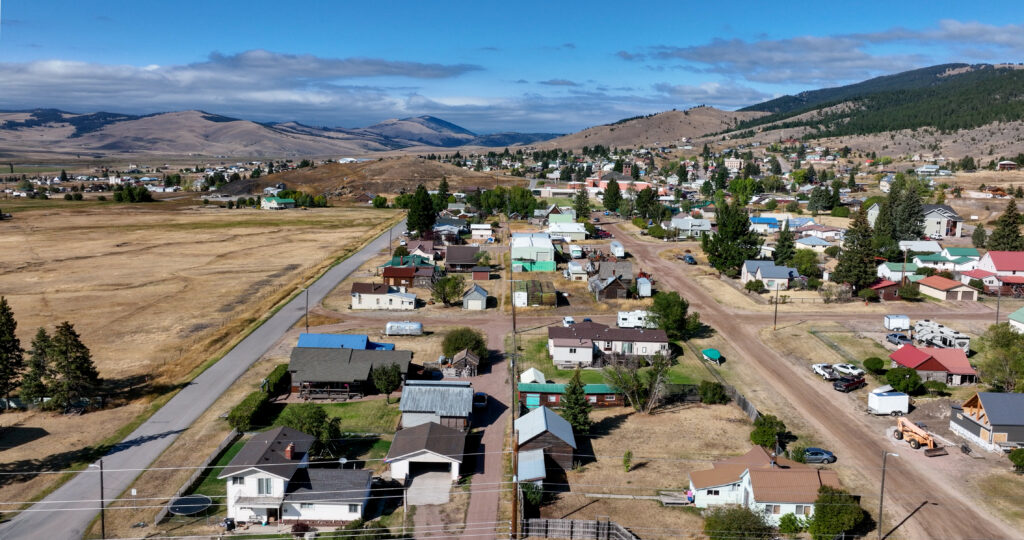 An aerial drone view of Philipsburg, Montana, showing a small historic town nestled among open ranchlands and rolling hills, with residential streets, colorful metal rooftops, scattered trees, and sweeping mountain ridgelines under a partly cloudy blue sky. Photo by Sarah Bolt, Tempest Technologies.