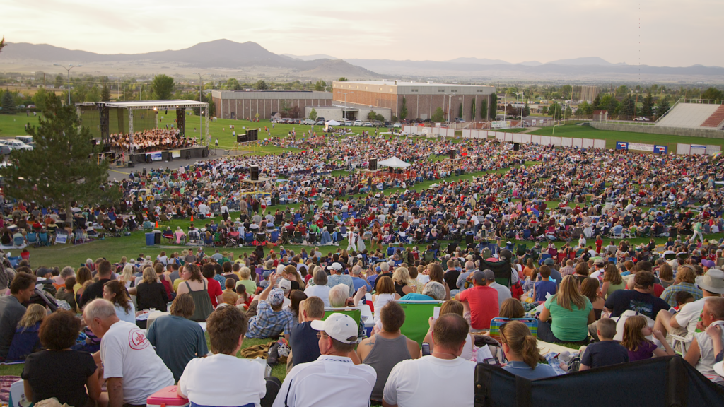 An overhead view of thousands of concert-goers seated on blankets and lawn chairs on the Carroll College hillside for Symphony Under the Stars in Helena, Montana, with the Helena Symphony on a lit outdoor stage and the surrounding valley and mountains fading into the evening light. Photo by Mike Dreesman, Tempest Technologies.