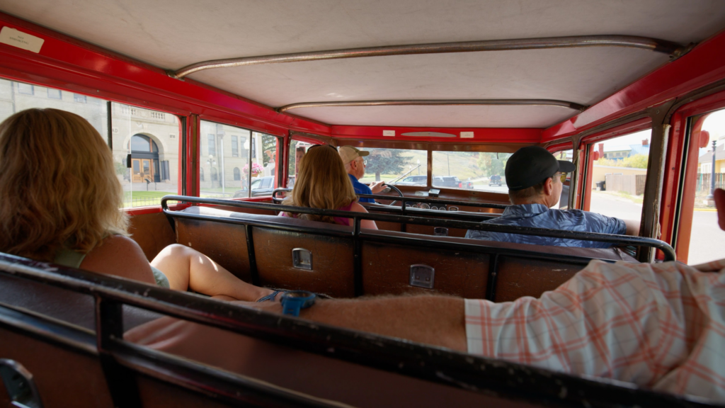 The view from the back seat inside the vintage Tour Anaconda red bus, looking forward toward the driver and two passengers seated on original brown leather bench seating, with the bus's panoramic windows framing a sunny summer pass through downtown Anaconda, Montana — the Deer Lodge County Courthouse visible through the windshield, capturing the immersive, old-school charm of a historic guided tour through one of Southwest Montana's most architecturally rich small towns.