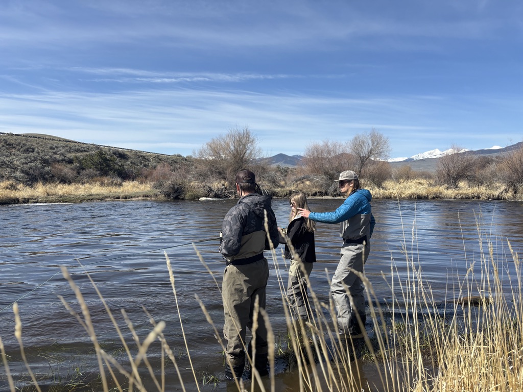 Three anglers standing in the shallows of the Big Hole River in Southwest Montana during a guided fly fishing trip in early winter, with a fishing guide in a blue jacket and waders appearing to a young angler while a second adult angler in camouflage waders stands nearby holding a fly rod, dry golden grass and dormant willow shrubs lining the near bank in the foreground, rolling sage covered hillsides rising behind the far bank, bare cottonwood trees stretching along the river corridor in the middle distance, and the snow capped Pioneer Mountains visible on the horizon under a wide blue Montana sky with wispy clouds, capturing the family friendly and guided fly fishing experience available on the Big Hole River near Twin Bridges, Montana and the Tony Schoonen Fishing Access Site in Southwest Montana.