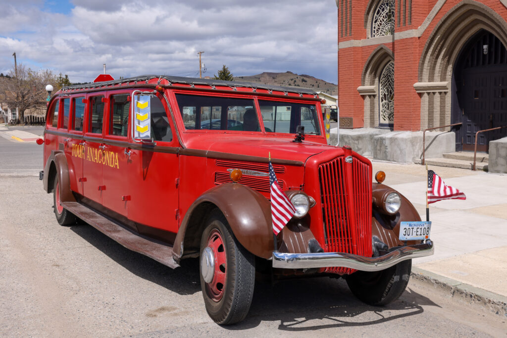 A vintage red and brown White Motor Company tour bus with "Tour Anaconda" lettered on the side, parked on a downtown Anaconda, Montana street beside a historic… A vintage red and brown White Motor Company tour bus with "Tour Anaconda" lettered on the side, parked on a downtown Anaconda, Montana street beside a historic red brick Gothic-style church, decorated with two small American flags on the front grille and featuring classic 1930s-era styling with chrome details, panoramic windows, and a Montana license plate — a charming nod to heritage tourism and small-town exploration in Southwest Montana's Deer Lodge County.