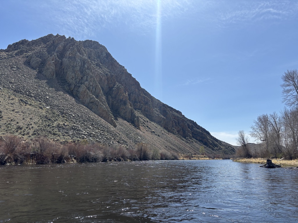 A wide view looking upstream along the Big Hole River at the Tony Schoonen Fishing Access Site near Twin Bridges, Montana, formerly known as Notch Bottom or The Notch to locals, with the towering rocky peak of the Hogback Ridge dominating the left side of the frame and rising steeply above the river corridor, its jagged exposed rock faces and sage covered slopes reflecting the ancient geology of the Quadrant Formation, a sandstone and quartzite formation pushed upward by tectonic forces over 75 million years ago, while the calm wide river flows through the narrow canyon carved over millions of years into the hard rock below, lined on the right bank by dormant red willow scrub, bare cottonwood trees, and dry spring grasses under a bright blue sky with wispy high clouds and a single ray of sunlight breaking through above the ridgeline, capturing the remote rugged and historically significant landscape of the lower Big Hole River in Southwest Montana in early spring.