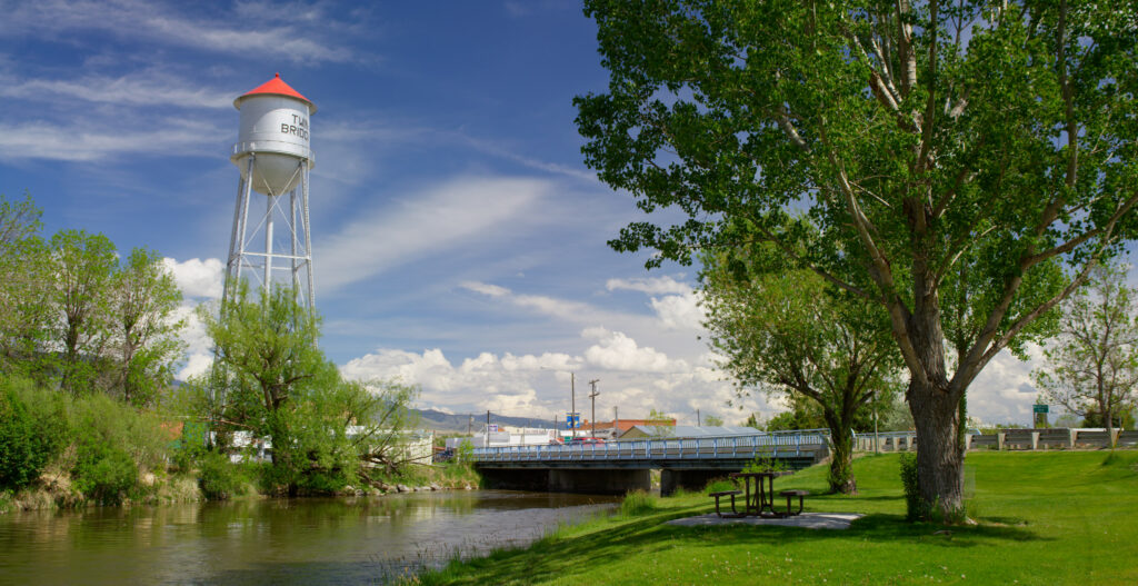 A scenic summer view of downtown Twin Bridges, Montana featuring the town's iconic white water tower with a red cap and Twin Bridges lettering rising above lush green trees on the left, a small river bridge with blue metal railings spanning what is believed to be the Beaverhead River in the center of the frame, a manicured green park with a picnic table and a large shade tree in the foreground on the right, and a wide Montana sky filled with dramatic billowing white clouds above the small town's rooftops and distant mountain ridgeline, capturing the charming small town character and riverside setting that makes Twin Bridges a popular base camp for fly fishing the Big Hole, Beaverhead, Ruby, and Jefferson Rivers in Southwest Montana.