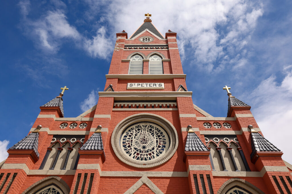 An upward-looking perspective of the red brick Gothic Revival facade of St. Peter's Catholic Church in Anaconda, Montana, showcasing the ornate rose window with intricate lacework stonework at center, twin flanking turrets topped with gold crosses, a tall central bell tower with arched windows and a gold cross finial, and decorative corbeling along the roofline — all set against a vivid blue sky with scattered clouds, highlighting the grandeur of Anaconda's historic Catholic heritage and copper boom-era architecture.