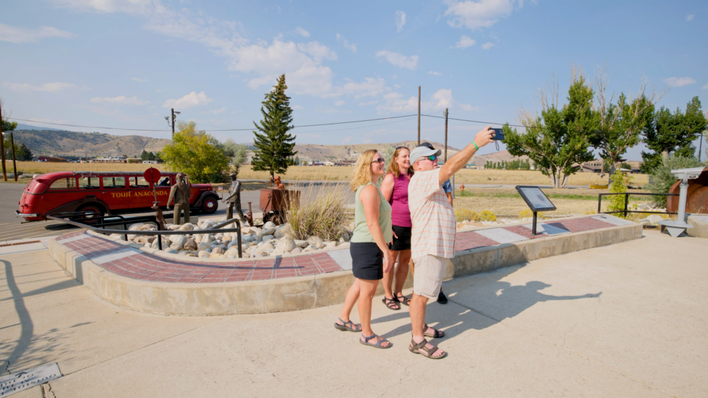 Three visitors taking a selfie together on a sunny summer day at Anaconda Stack State Park in Anaconda, Montana, with the vintage red Tour Anaconda bus and a bronze miner sculpture visible in the background alongside interpretive signage and a decorative river rock plaza — the surrounding open grasslands and foothills of Southwest Montana stretching out under a partly cloudy blue sky, capturing the welcoming, story-rich experience of heritage tourism in Deer Lodge County.