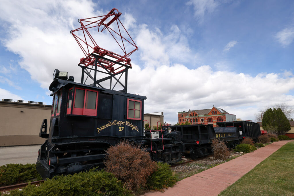 A preserved Anaconda Copper Mining Company electric locomotive — engine number 97 — displayed in black and gold livery on a short stretch of track outside the Discover Anaconda visitor's center, with a red overhead pantograph assembly raised above the cab and a Butte, Anaconda & Pacific Railway ore car numbered 451 coupled behind it, set against a partly cloudy sky with a historic red brick downtown Anaconda building visible in the background, capturing the copper smelting and industrial railroad heritage that defined Anaconda, Montana as one of the most important mining and smelting cities in the American West.