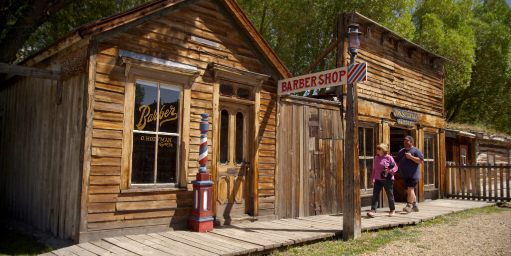 Two visitors walking along a weathered wooden boardwalk past a historic 19th-century barber shop building with a red white and blue barber pole, hand-painted signage reading C. Hoffman Proprietor, and a Jos. Sullivan Saddlery sign next door, along the preserved frontier-era main street of Virginia City Montana on a sunny summer day. Photo by Tempest Technologies.