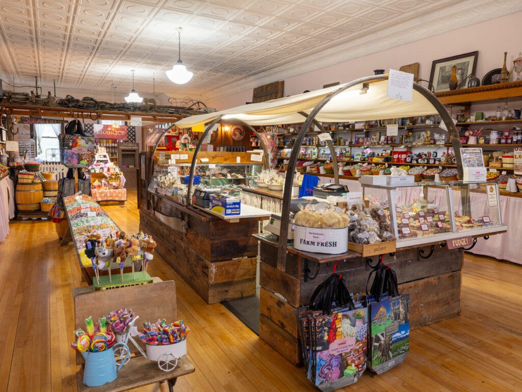 The charming interior of Cousin's Candy Shop in Virginia City, Montana, a must-stop on any Southwest Montana road trip. A large arched glass display case anchors the center of the room, stocked with fudge, huckleberry bark, and homemade baked goods. Rustic wooden barrels and long candy counters line the walls, filled with bulk sweets, licorice, and novelty candies. Colorful lollipops and stuffed animal pops are arranged near the entrance, and Montana-themed tote bags hang from the display case. An original ornate pressed tin ceiling and warm pendant lighting add to the historic atmosphere of this beloved Virginia City sweet shop. Photo by Sarah Bolt, Tempest Technologies.