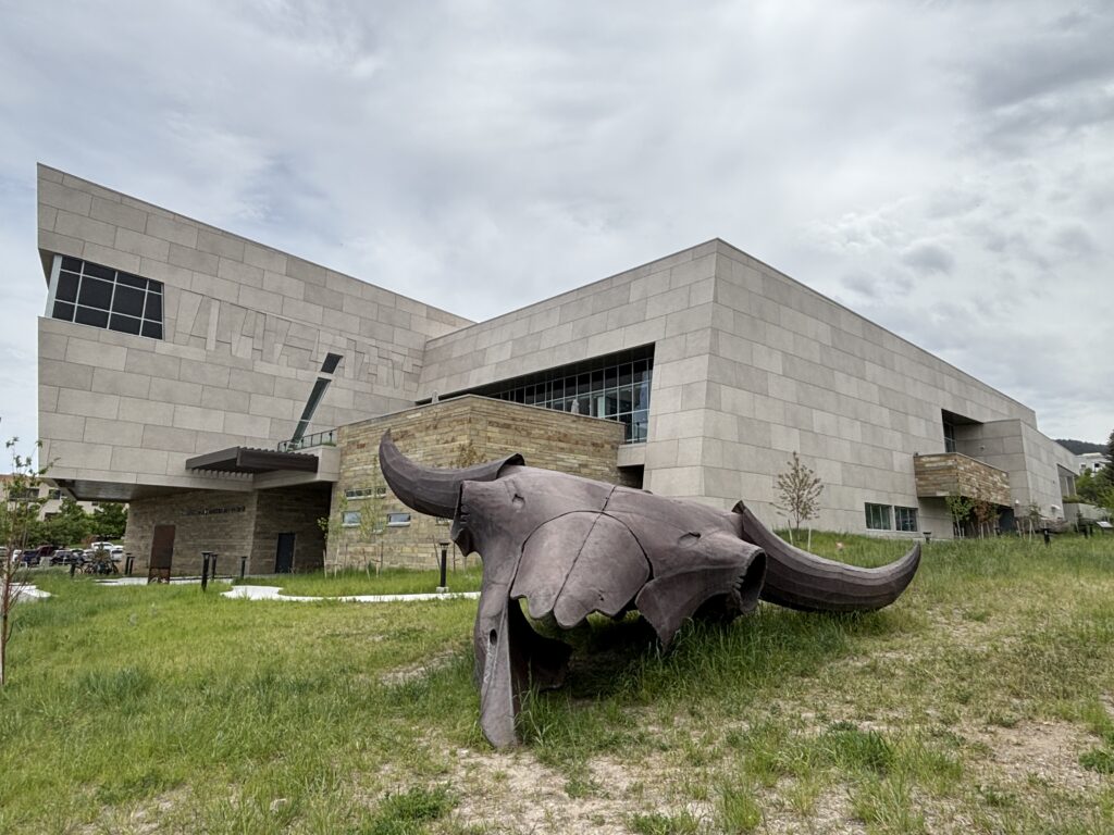Exterior of the Montana Heritage Center in Helena, Montana, home of the Montana Historical Society. A massive bronze bison skull sculpture rests in native grasses in the foreground, its curved horns stretching toward the museum's modern limestone facade. The angular building features pale stone walls, warm stacked-rock accents, and large windows beneath an overcast Montana sky. The Montana Heritage Center hosts the Montana History Fest in Helena, June 24–28. Photo by Alyssa Starr, Tempest Technologies.
