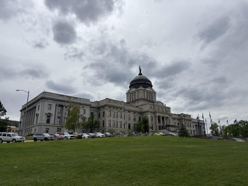 The Montana State Capitol in Helena, Montana, viewed from across the green Capitol lawn, with the copper dome and the "Montana" statue rising against a dramatic cloudy sky. The granite building's columned facade and rows of state and tribal flags line the entrance. The Capitol lawn hosts the Alive at 5 concert during the Montana History Fest in Helena for the first time. Alive at Five is a free, public, outdoor concert series in Helena, Montana. Photo by Alyssa Starr, Tempest Technologies.