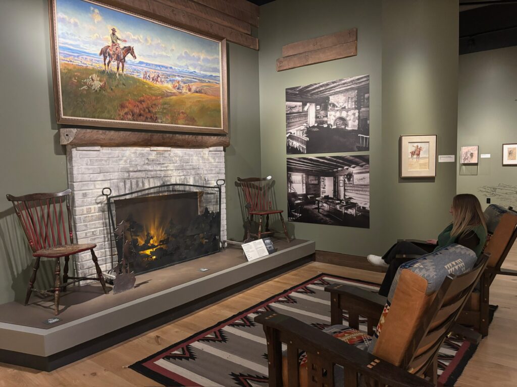 A guest relaxes in a leather and Pendleton-style armchair inside the Charles M. Russell Gallery at the Montana Heritage Center in Helena, Montana. The gallery recreates the cozy feel of Russell's home, with a whitewashed brick fireplace, antique Windsor chairs, a Navajo-style rug, and historic black-and-white photographs of the artist's log cabin studio. Above the mantel, one of Russell's western paintings shows a cowboy on horseback overlooking a sweeping prairie. Photo by Alyssa Starr, Tempest Technologies.