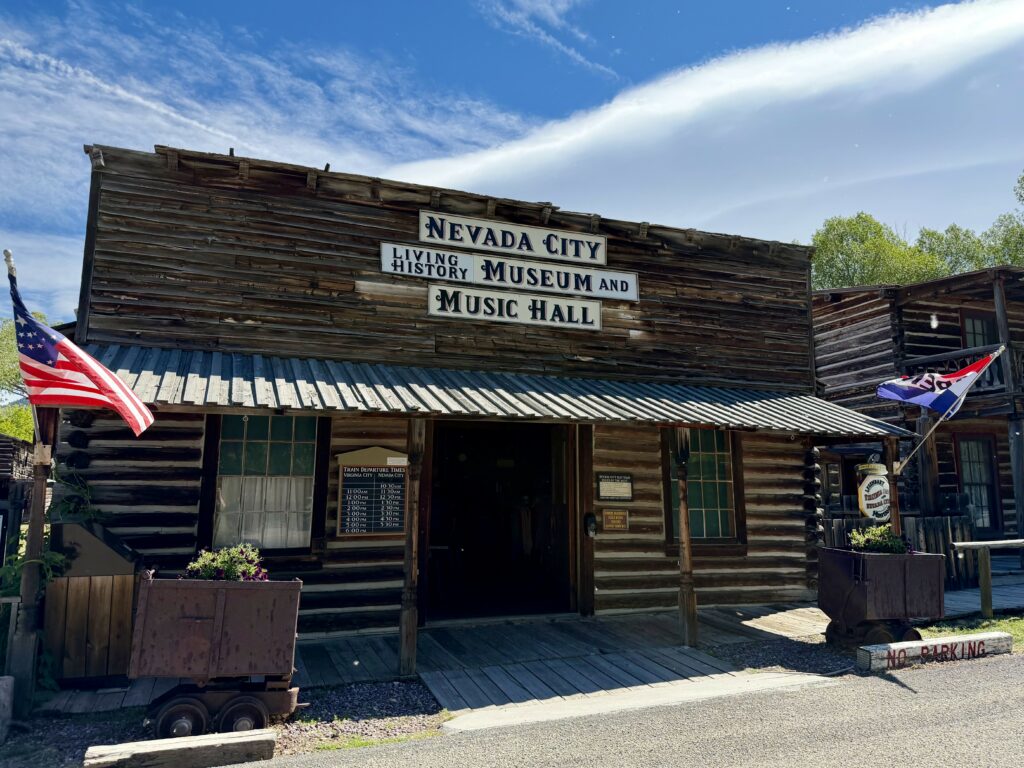 The exterior of the Nevada City Living History Museum and Music Hall in Nevada City, Montana. The rustic log-and-wood-plank building displays a large painted sign across the facade. An American flag flies from the left side of the covered porch, and a vintage mining ore cart used as a planter sits in the foreground. A train departure schedule board listing Virginia City and Nevada City times is visible near the entrance. Blue skies and green trees frame the building, evoking the preserved Gold Rush-era atmosphere of this Southwest Montana ghost town.
