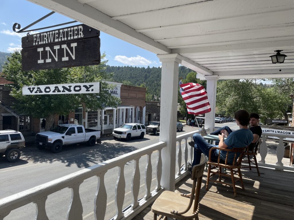 Two visitors relax in wooden rocking chairs on the covered second-story porch of the Fairweather Inn in Virginia City, Montana, taking in a sweeping view of the town's historic main street. The inn's rustic wooden sign and a Vacancy placard hang in the foreground. Across the street, preserved 19th-century storefronts — including a steakhouse and a mail and mercantile building — line the block, with an American flag waving and tree-covered hills rising in the background. A Blacksmith and Wagon sign is partially visible to the right. Photo by Sarah Bolt, Tempest Technologies.