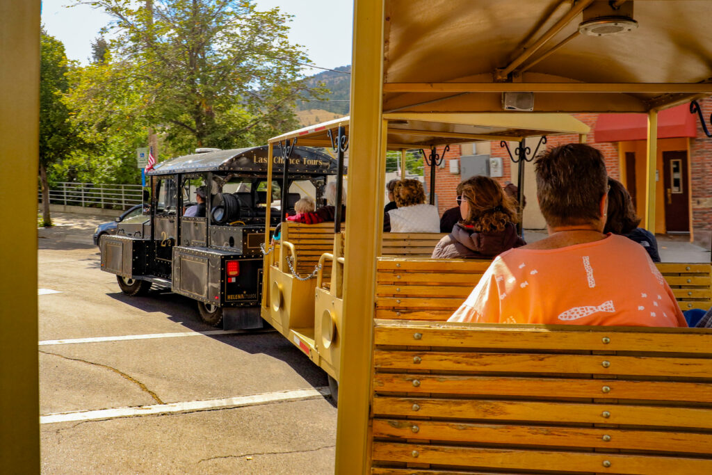Riders aboard the Last Chance Tour Train in Helena, Montana, photographed from a wooden bench seat as the open-air tram rounds a corner. The black engine, marked "Last Chance Tours" and flying an American flag, pulls passenger cars filled with visitors past brick buildings and tree-lined streets, with the hills above Helena in the background. The hour-long tour winds through Helena's historic districts, including Last Chance Gulch and Reeder's Alley. Photo by Sarah Bolt, Tempest Technologies.