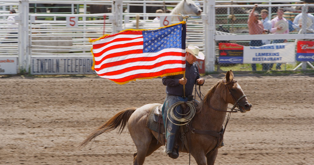 2:12 PM A cowboy in a white hat and blue shirt rides a sorrel horse across a dirt arena carrying a large American flag during the grand entry at the Lincoln Rodeo in Lincoln, Montana. The flag streams behind him as he lopes past the chutes, numbered gates, and a crowd of spectators visible in the background alongside sponsor signage. The Lincoln Rodeo is one of Southwest Montana's beloved small-town summer traditions, drawing locals and visitors alike for a classic Western celebration complete with traditional rodeo events, community atmosphere, and Montana ranch culture. Photo by Mike Dreesman, Tempest Technologies.