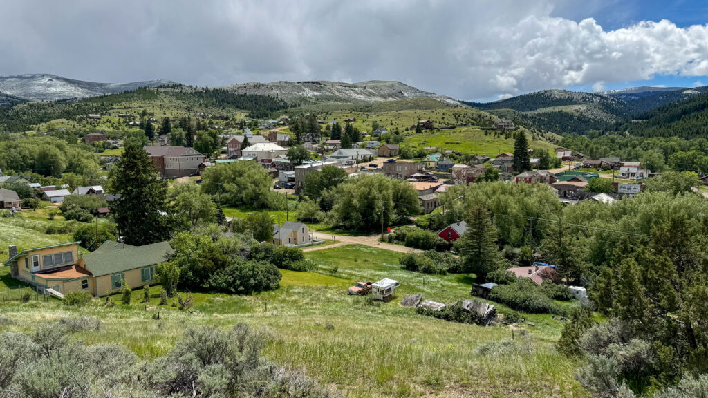 An elevated panoramic view of Virginia City, Montana, as seen from the vintage fire truck tour, revealing the full sweep of this historic gold rush town tucked into a mountain valley. Historic storefronts, a red brick courthouse, and a mix of 19th-century and early 20th-century buildings are visible among the trees, with lush green hillsides and snow-dusted peaks of the Tobacco Root Mountains rising in the background. An Overland Mail sign is visible on the right side of the frame. This sweeping perspective captures why Virginia City, a Montana National Historic Landmark, remains one of the most scenic and well-preserved small towns in Southwest Montana. Photo by Sarah Bolt, Tempest Technologies.