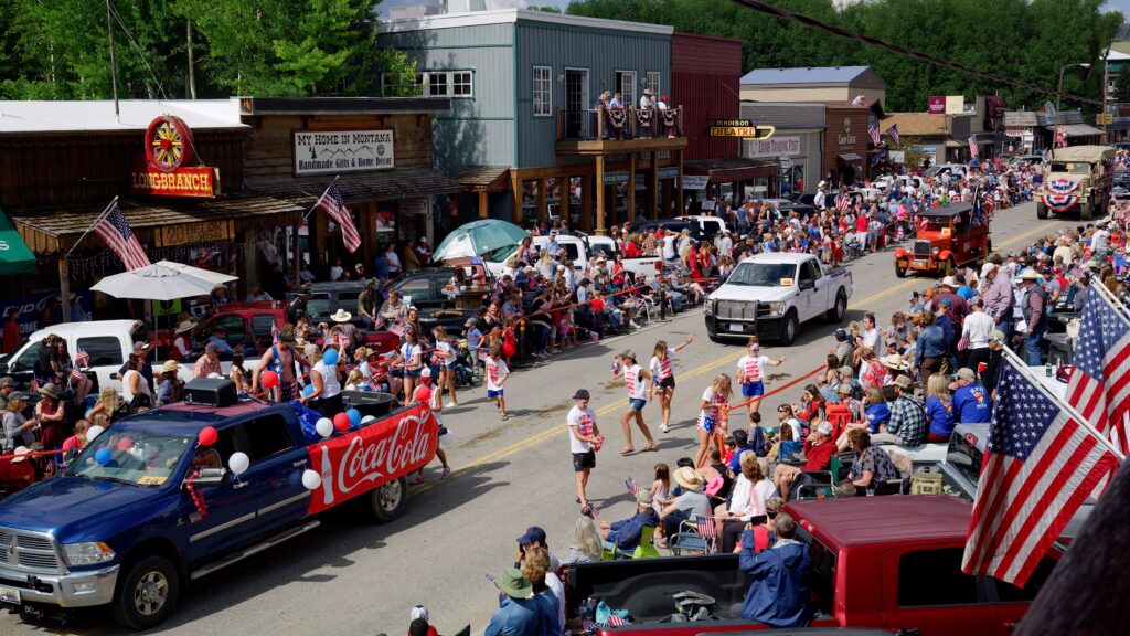 A massive crowd lines both sides of Main Street in Ennis, Montana, for the town's Fourth of July parade on a bright summer day. Decorated trucks, floats, and parade participants fill the street as children in red, white, and blue dart between vehicles tossing candy to spectators. A Coca-Cola sponsored truck with red and blue balloons rolls through the parade route while onlookers wave American flags from lawn chairs, truck beds, and a second-floor balcony overlooking the festivities. Historic storefronts including the Longbranch, My Home in Montana, the Madison Theatre, and Ennis Trading Post line the route, packed with locals and visitors celebrating the holiday. The Ennis Fourth of July parade is one of Southwest Montana's most beloved small-town Independence Day traditions, drawing crowds from across the region to the Madison Valley for a classic Montana summer celebration complete with live music, rodeo, and community spirit. Photo by Mike Dreesman, Tempest Technologies.