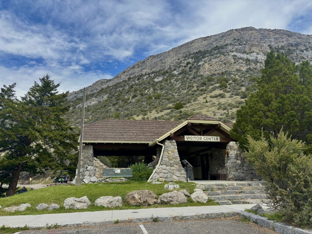 The stone and timber Cave Visitor Center at Lewis and Clark Caverns State Park sits at the base of a rocky mountain slope, with a stone staircase leading up to the entrance and a covered pavilion structure to the left surrounded by pine trees and boulders.