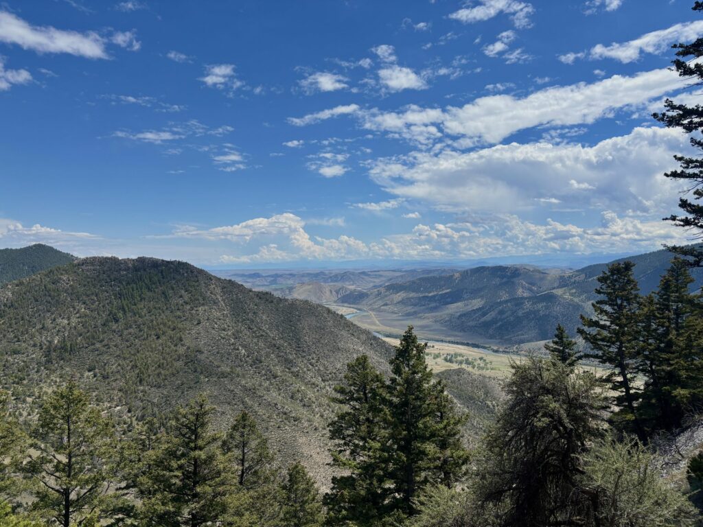 A sweeping view of the Jefferson River Canyon from the cave entrance on the Classic Tour at Lewis and Clark Caverns State Park, showing forested ridgelines, the winding river valley floor, and distant mountains beneath a wide blue sky dotted with clouds.